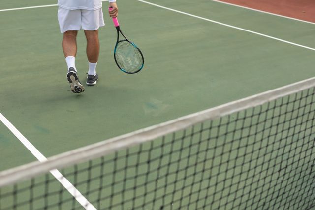 Low section of Caucasian man wearing tennis whites spending time on a court playing tennis on a sunny day, holding a tennis racket. Hobby sport leisure time.