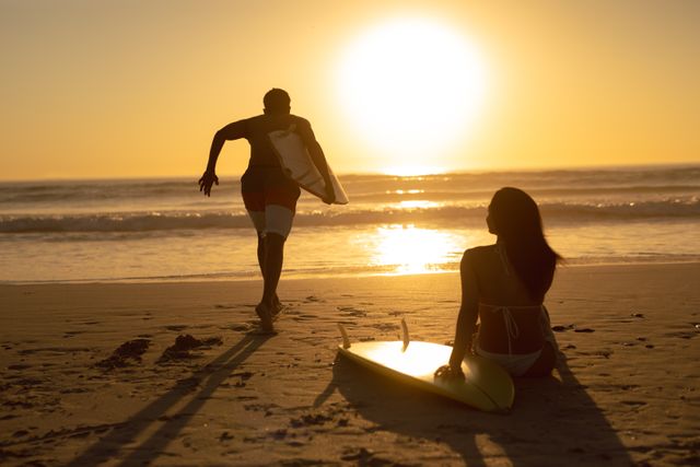 Man running with surfboard while woman relaxing on the beach during sunset - Download Free Stock Photos Pikwizard.com