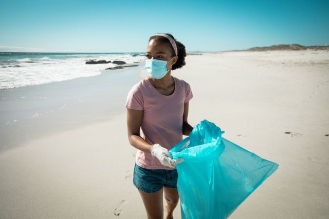 African american woman wearing face mask and gloves putting rubbish in refuse sack on sunny beach - Download Free Stock Photos Pikwizard.com