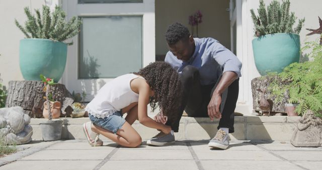 Happy african american father sitting on garden step with daughter tying his shoe. Fatherhood, childhood, care, togetherness and domestic life.