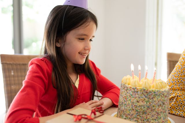 Little caucasian girl wearing a birthday hat sitting on the dinner table in front of a birthday cake with four candles. sitting beside her is her mom.