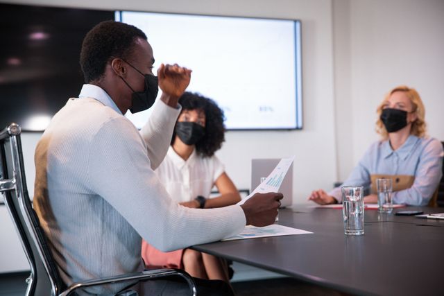African american businessman and female colleagues, wearing face masks and talking in meeting room. working in business at a modern office during coronavirus covid 19 pandemic.