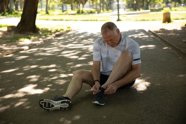 Senior Caucasian man working out in the park wearing sports clothes, sitting on path tying shoelaces. Retirement healthy lifestyle activity.