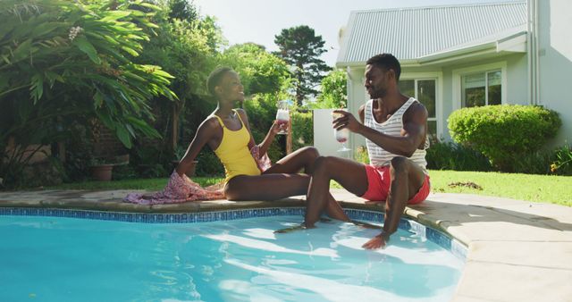 Happy african american couple sitting by swimming pool making a toast with drinks in sunny garden. staying at home in isolation during quarantine lockdown.