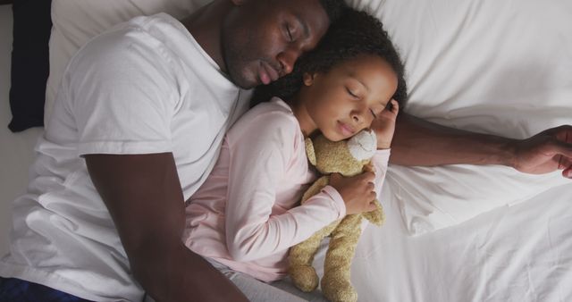 Happy african american father lying asleep in bed beside daughter holding teddy bear. Fatherhood, childhood, care and domestic life.