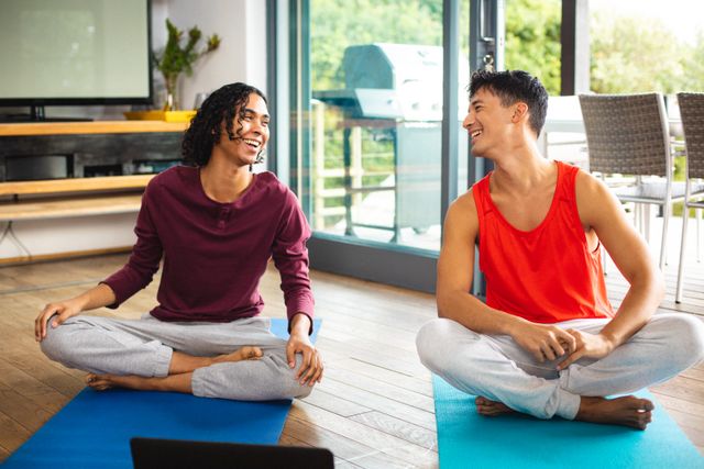 Multiracial gay couple looking at each other and laughing while meditating on mats at home - Download Free Stock Photos Pikwizard.com