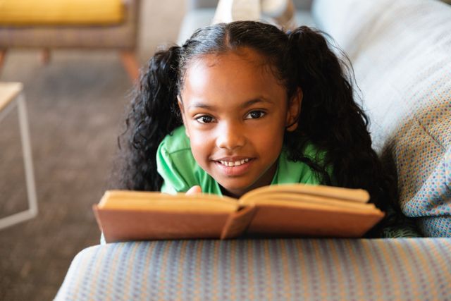 Portrait of smiling african american elementary girl with book lying on sofa in school play room - Download Free Stock Photos Pikwizard.com