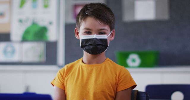 Portrait of mixed race schoolboy wearing face mask, sitting in classroom looking at camera. children in primary school during coronavirus covid 19 pandemic.