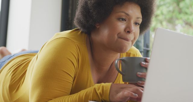 Image of happy plus size african american woman lying on floor with coffee and using laptop. lifestyle, leisure, spending free time at home with technology.