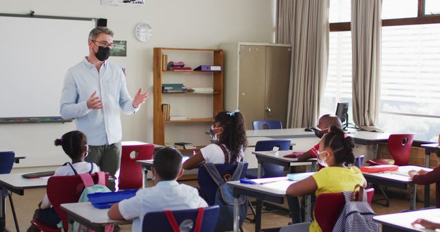 Caucasian male teacher wearing face mask teaching students in the class at school. hygiene and social distancing at school during covid 19 pandemic.