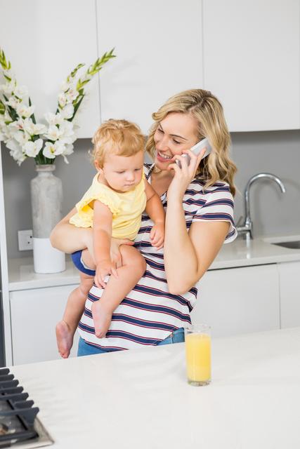 Mother talking on mobile phone while holding her baby girl in kitchen - Download Free Stock Photos Pikwizard.com