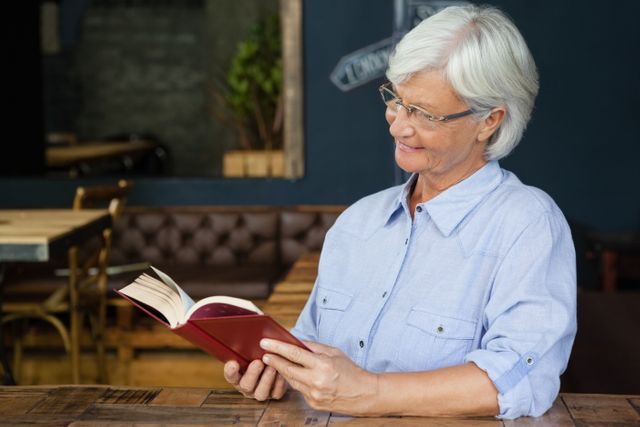 Smiling senior woman reading book while sitting by table - Download Free Stock Photos Pikwizard.com