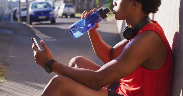 Fit african american man exercising in city taking a break, using smartphone, sitting drinking water. fitness and active urban outdoor lifestyle.