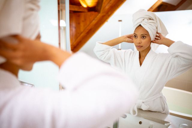Mixed race woman wearing robe and towel turban, looking in mirror - Download Free Stock Photos Pikwizard.com