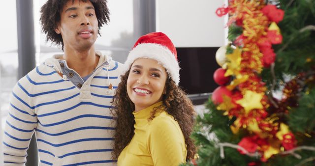 Image of happy diverse couple in reindeer antlers and santa hat admiring christmas tree at home. Christmas, celebration, happiness, tradition and inclusivity concept.