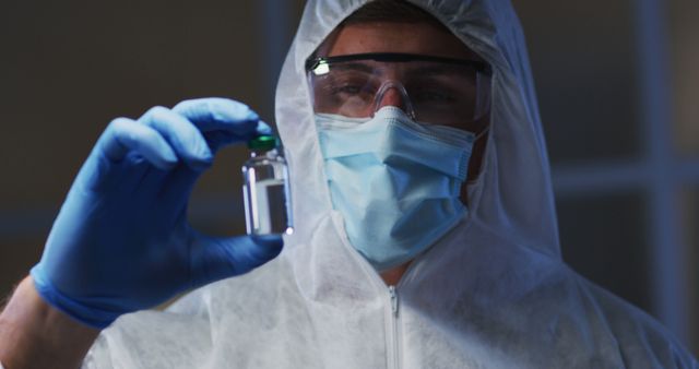Caucasian male medical worker wearing protective clothing mask and gloves holding vaccine vial in lab. healthcare, medical research technology and hygiene during coronavirus covid 19 pandemic.