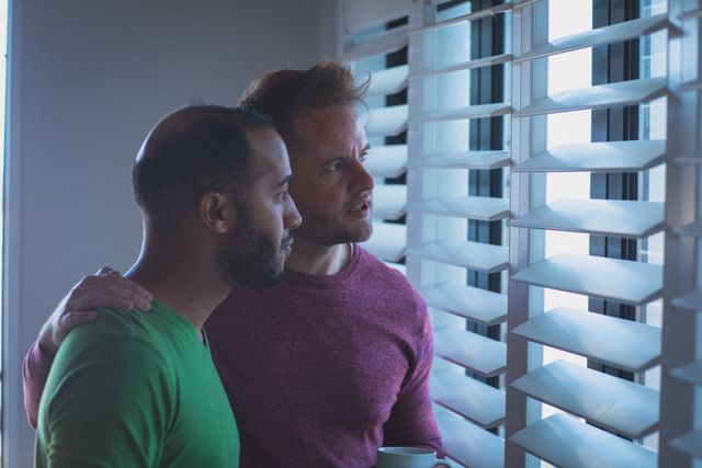 Diverse gay male couple with arms around each other looking out of window through blinds. staying at home in isolation during quarantine lockdown.
