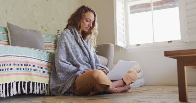 Smiling caucasian woman sitting on floor wearing blanket using laptop in sunny cottage living room. simple living in an off the grid rural home.