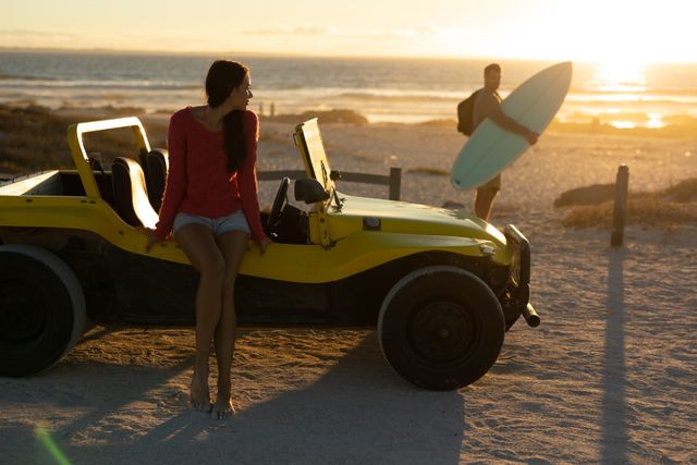 Woman sitting on beach buggy by sea at sunset, watching man carrying surfboard - Download Free Stock Photos Pikwizard.com