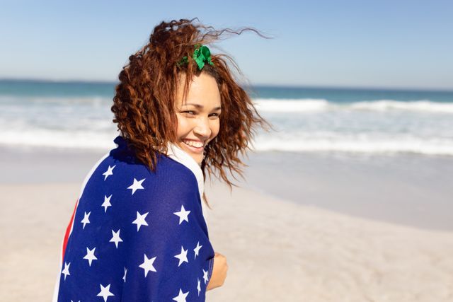 Rear view of beautiful happy young Mixed-race woman wrapped in american flag looking at camera on beach in the sunshine