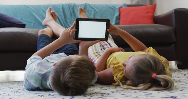 Happy caucasian brother and sister lying on floor in living room using tablet, copy space on screen. happy family, free time at home.