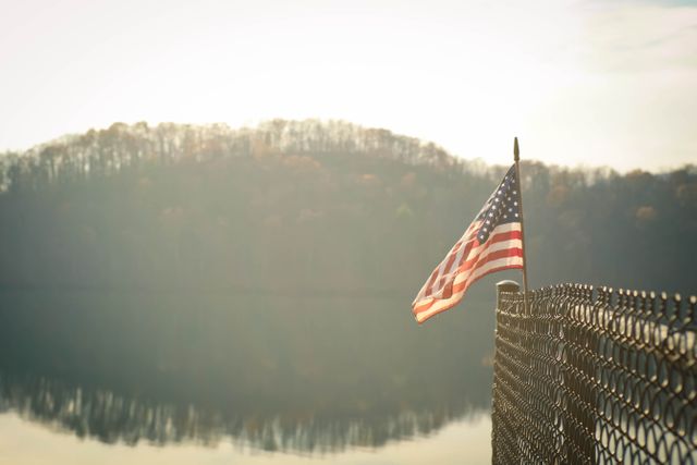American Flag on Edge of Fence Near Body of Water With Tree Silhouette in the Background Under White Sky - Download Free Stock Photos Pikwizard.com