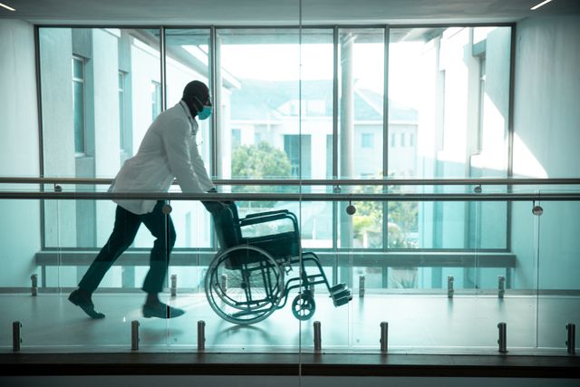 African american male doctor wearing face mask pushing wheelchair in hospital corridor - Download Free Stock Photos Pikwizard.com