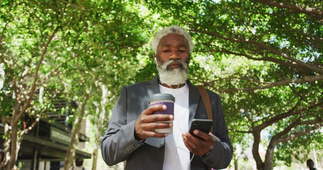 African american senior man holding smartphone drinking coffee while walking in corporate park. active senior lifestyle living concept