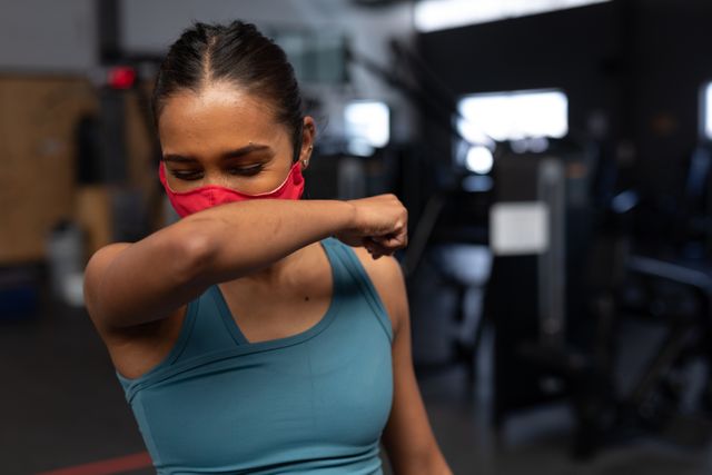 Caucasian woman covering her mouth and nose with her arm as she sneezes. she is wearing a red facemask while she is inside the gym