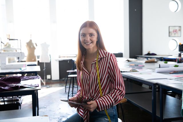 Front view of a young red haired Caucasian female fashion student with a yellow tape measure round her neck in a studio at fashion college, looking straight to the camera, smiling, holding computer tablet.