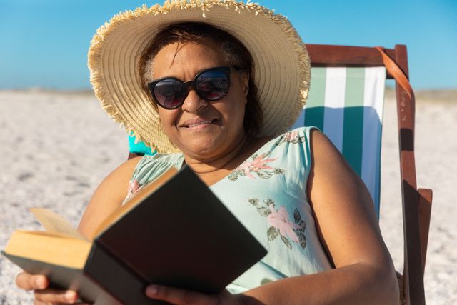 Smiling african american retired senior woman reading book sitting on folding chair at beach. unaltered, hobbies, active lifestyle, enjoyment and holiday concept.