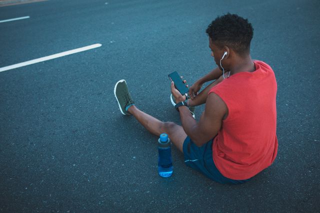 Fit african american man sitting on coastal road using smartphone and headphones - Download Free Stock Photos Pikwizard.com