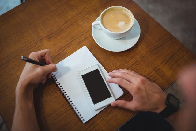 Man writing on notepad while using mobile phone in cafÃ©
