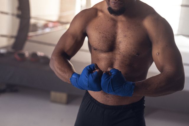 Midsection of shirtless macho african american young male boxer wearing blue wraps flexing muscles. Health club, unaltered, boxing, sport, training, strength and fitness concept.