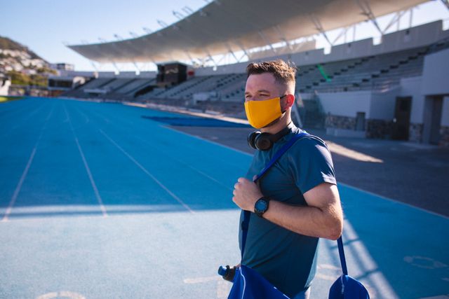 Caucasian male athlete wearing face mask, headphones and bag, standing on stadium - Download Free Stock Photos Pikwizard.com