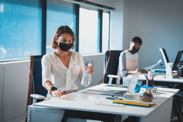 Two diverse businesswomen wearing face masks sitting at office desks, one disinfecting her workspace - Download Free Stock Photos Pikwizard.com