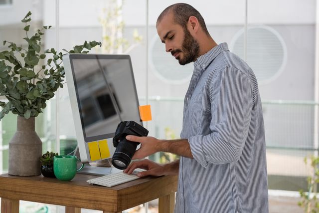 Designer examining camera while standing by computer - Download Free Stock Photos Pikwizard.com