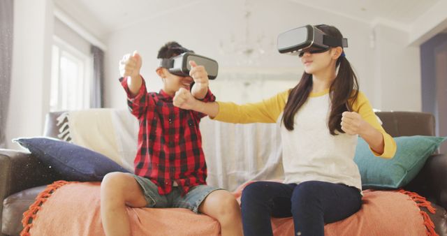 Happy asian brother and sister at home, sitting on couch in living room wearing vr headsets. at home in isolation during quarantine lockdown.