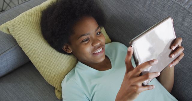 Happy african american teenage girl lying on sofa, using tablet. domestic lifestyle, using communication technology at home.