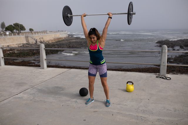 Front view of a strong Caucasian woman with long dark hair wearing sportswear exercising outdoors by the seaside on a sunny day, strength training lifting barbells above her head, concentrating, ball and kettlebell behind her.