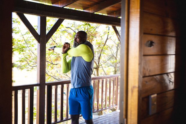 Bald african american senior man stretching hands while exercising in balcony seen through doorway. Log cabin, unaltered, vacation, beard, retirement, solitude, yoga, fitness and active lifestyle.