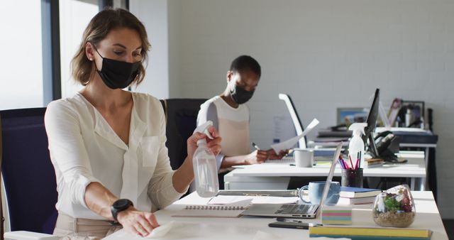 Two diverse female colleagues wearing face mask, sanitizing desk in office. independent creative business at a modern office during coronavirus covid 19 pandemic.