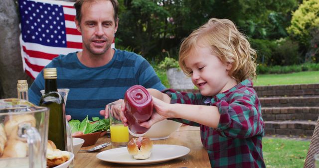 Smiling caucasian man watching son using ketchup during celebration meal in garden. family celebrating independence day eating outdoors together.
