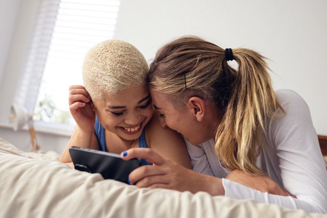 Happy multiracial young lesbian couple laughing while watching video over cellphone and lying on bed. Technology, cheerful, unaltered, love, togetherness, homosexual, lifestyle and home concept.