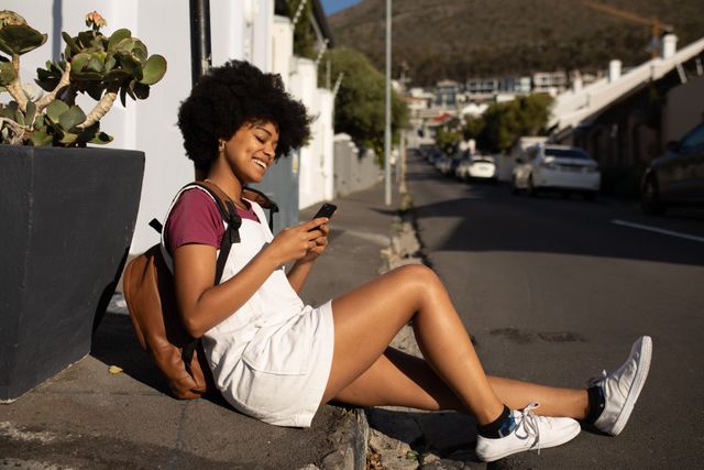 SIde view of a happy mixed race woman enjoying free time in a city on a sunny day, using a smartphone, sitting on a pavement, wearing white mini dress and backpack.