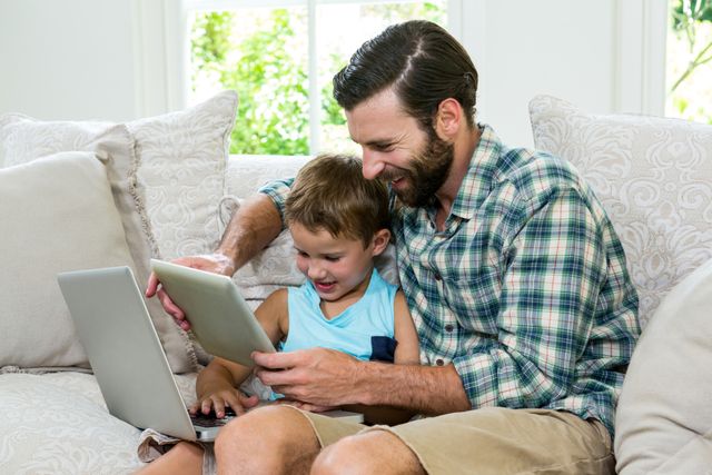 Playful son and father with laptop and digital tablet on sofa at home