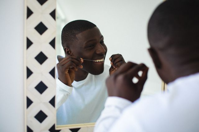 African american young man flossing his teeth while looking in mirror in bathroom, copy space - Download Free Stock Photos Pikwizard.com