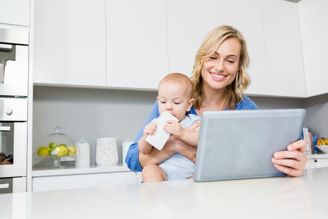 Mother holding baby boy while using digital tablet in kitchen - Download Free Stock Photos Pikwizard.com