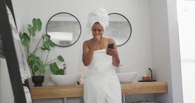 Happy african american woman brushing teeth and using smartphone in bathroom. domestic lifestyle, spending free time at home.