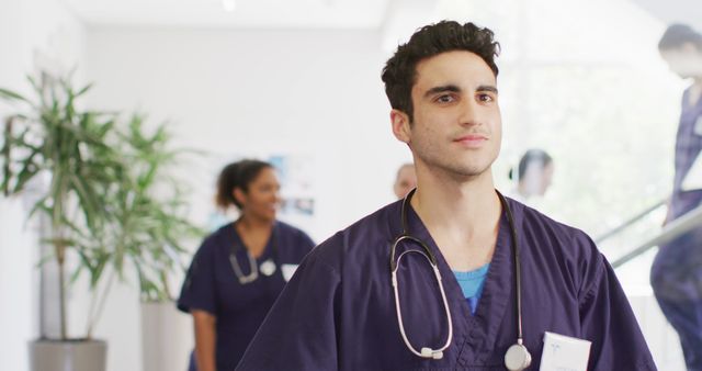 Image of smiling biracial male doctor walking in busy hospital corridor, with copy space. Hospital, medical and healthcare services.